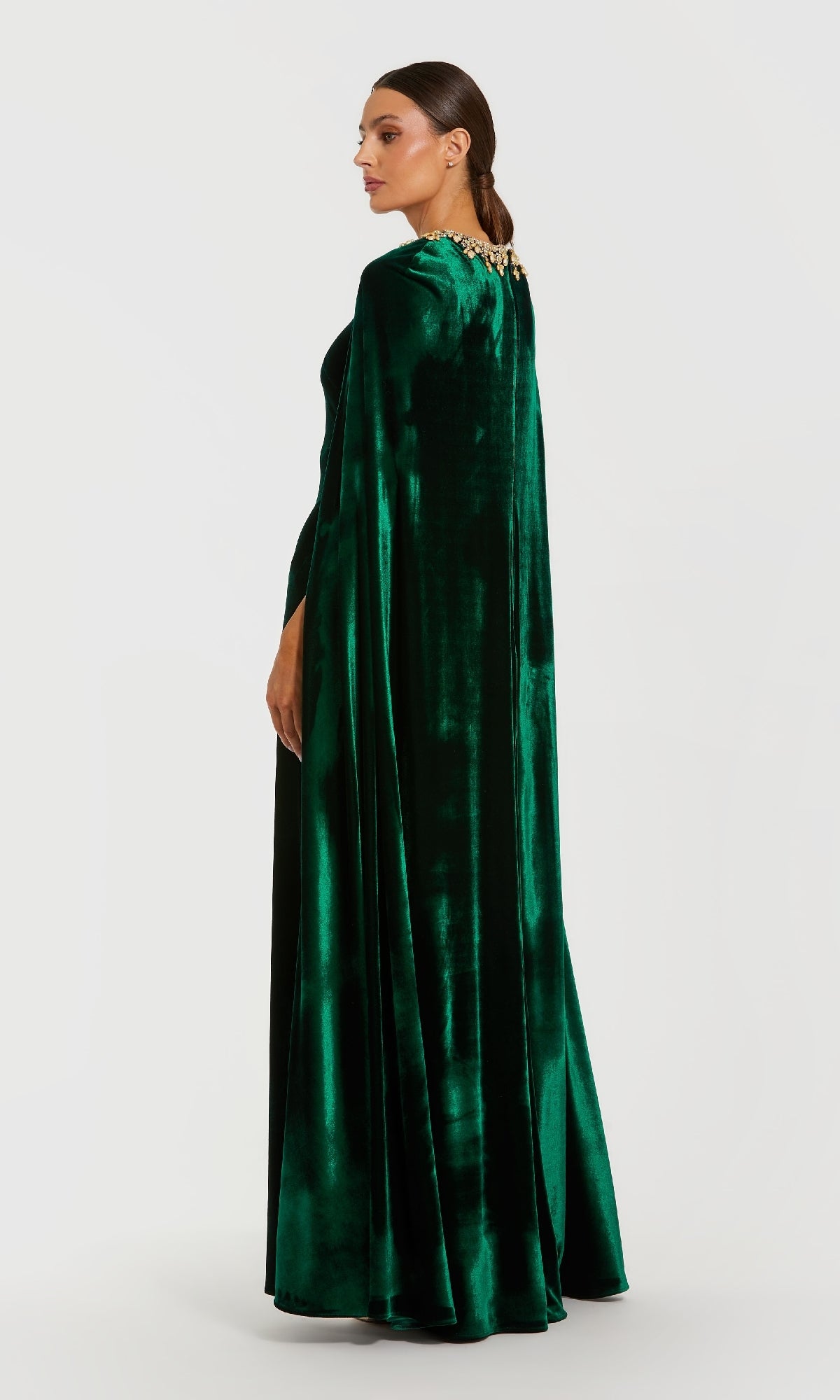 A woman stands in profile wearing the Green Velvet Caped Formal Dress: Mac Duggal 68977, a floor-length gown with a cape and gold embellishments at the neckline.
